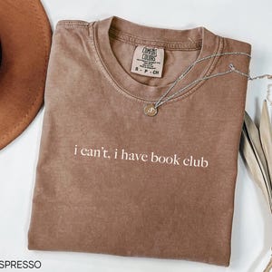 May include: A brown t-shirt with the text "i can't, i have book club" printed in white. The shirt is made of a soft, comfortable material and has a relaxed fit.