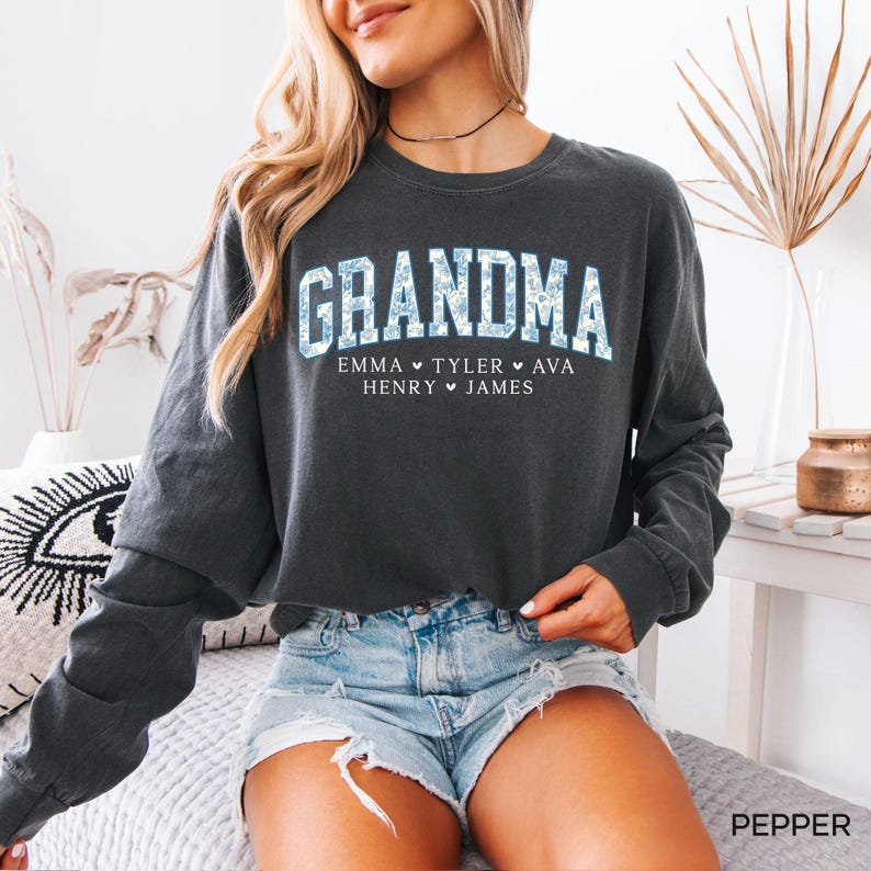 May include: Dark gray long-sleeve sweatshirt with the word "GRANDMA" in large, light blue floral print letters. Below the word are the names Emma, Tyler, Ava, Henry, and James. The sweatshirt is worn with denim shorts.