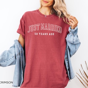 May include: Crimson-colored t-shirt with the text "JUST MARRIED" above "50 YEARS AGO" in white lettering. The shirt has short sleeves and a crew neck. A light blue denim jacket is draped over the shoulders.