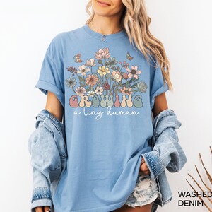 May include: Light blue t-shirt with a floral design and the text "Growing a tiny human" in pastel colors. The shirt has rolled-up sleeves and a relaxed fit. Washed denim.