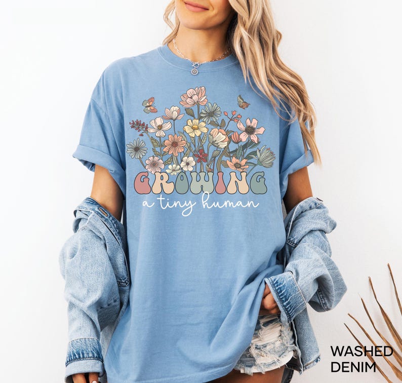 May include: Light blue t-shirt with a floral design and the text "Growing a tiny human" in pastel colors. The shirt has rolled-up sleeves and a relaxed fit. Washed denim.