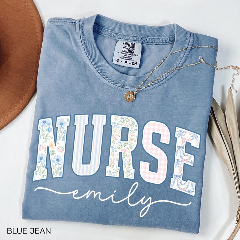 May include: Blue jean colored t-shirt with the word "NURSE" in floral and striped patterns, and the name "emily" in a cursive font. A gold necklace with a pendant rests on the shirt.