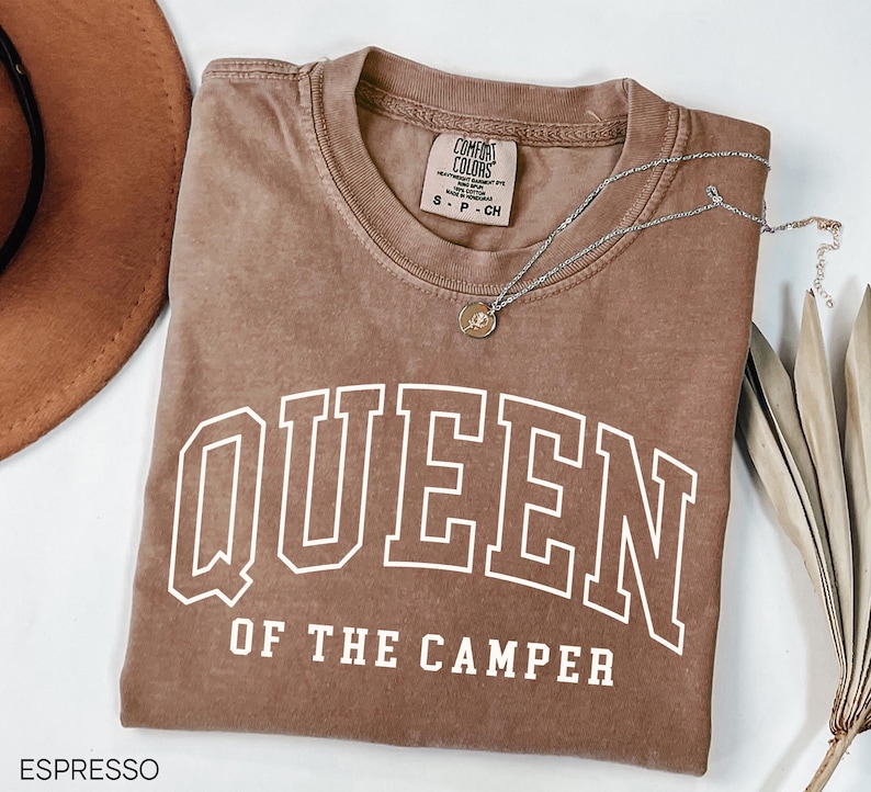 May include: A brown t-shirt with the text "QUEEN OF THE CAMPER" in white. The shirt has a distressed look and is made by Comfort Colors.