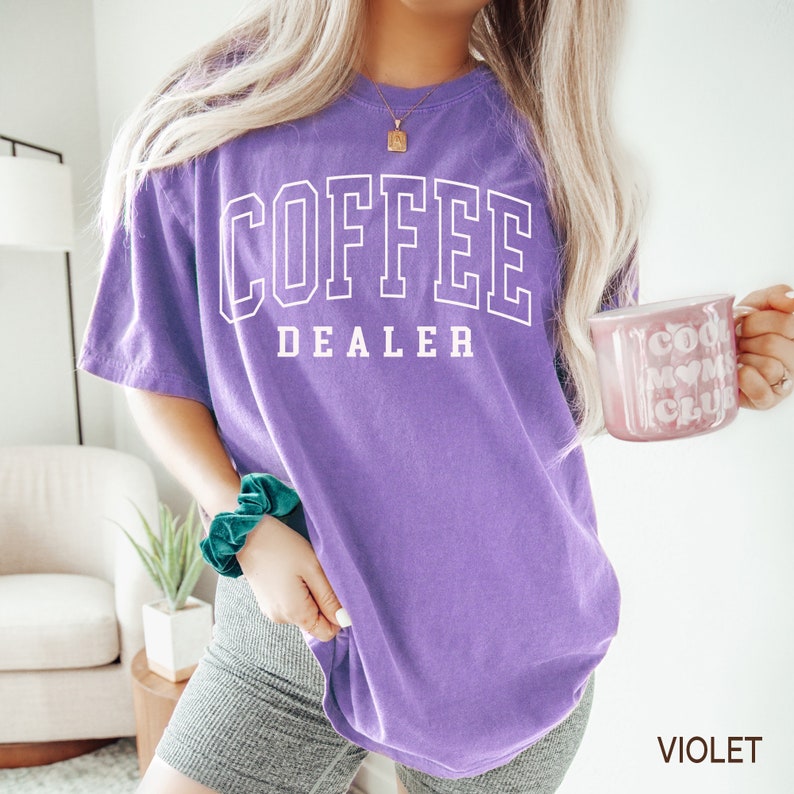 May include: A violet-colored t-shirt with the words "COFFEE DEALER" in white block letters. The shirt is a casual style, and the text has a collegiate font. The person is holding a pink mug with the words "COOL MOM CLUB".