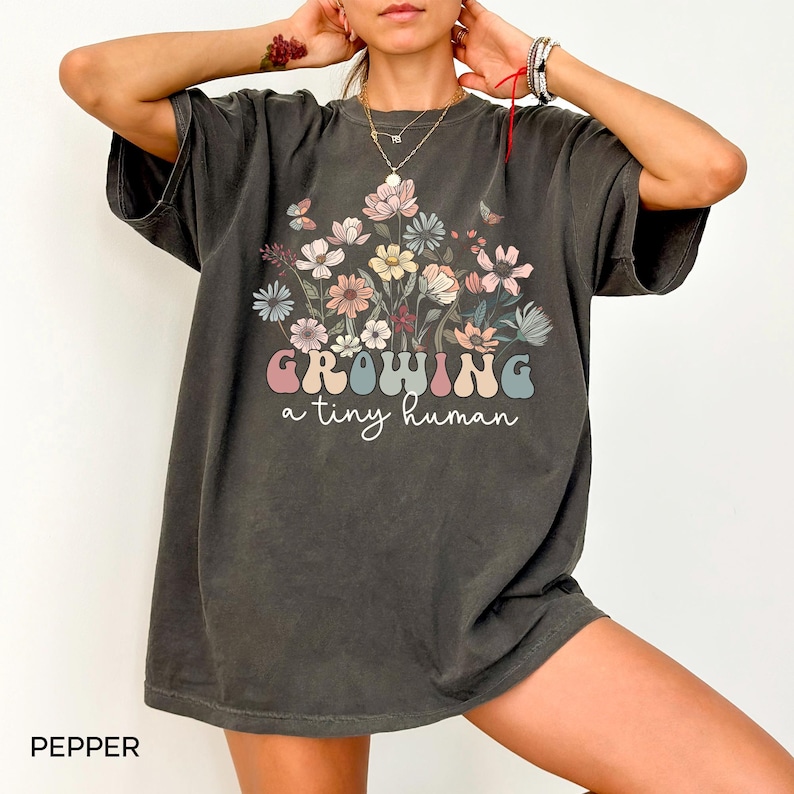 May include: Dark grey t-shirt with a floral design and the text "GROWING a tiny human" in pastel colors. The shirt features a crew neck and short sleeves. The word "PEPPER" is printed at the bottom left corner.