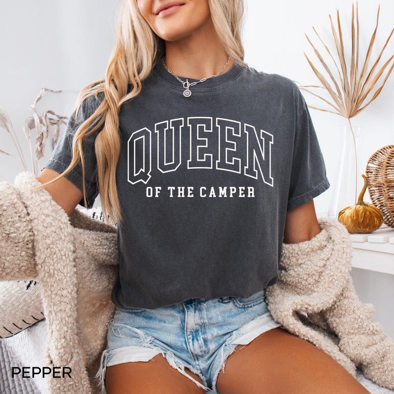 May include: A dark gray t-shirt with the text "QUEEN OF THE CAMPER" in white. The shirt is worn by a person with long blonde hair and is styled with denim shorts.