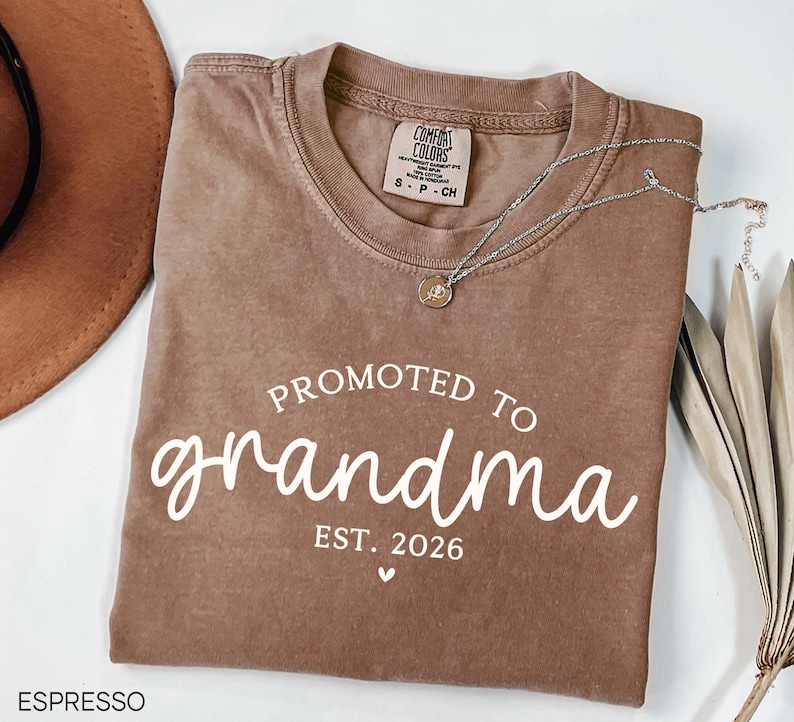 May include: Espresso-colored t-shirt with the text "PROMOTED TO grandma EST. 2026" in white script. A silver necklace with a pendant rests on the shirt. A brown hat and dried plant are in the background. The shirt is a Comfort Colors brand.