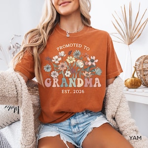 May include: Burnt orange t-shirt with the text "Promoted to Grandma Est. 2026" in a floral design. The shirt has a vintage look and is made of a soft material. The design features pastel colors.