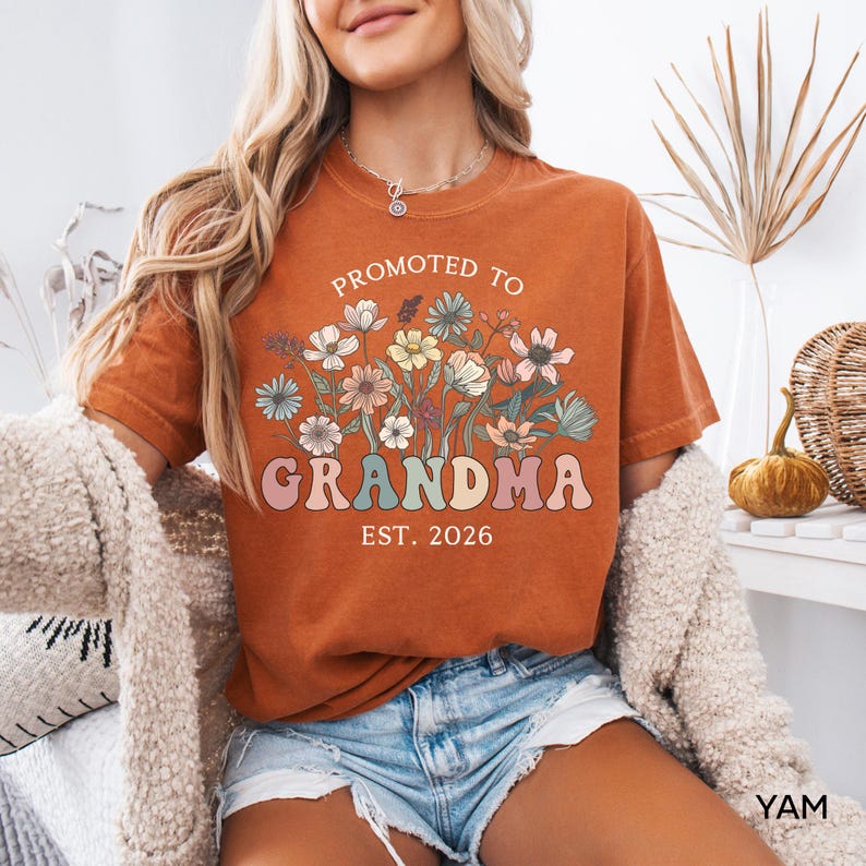 May include: Burnt orange t-shirt with the text "Promoted to Grandma Est. 2026" in a floral design. The shirt has a vintage look and is made of a soft material. The design features pastel colors.