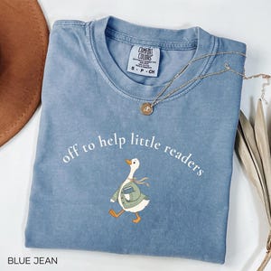 May include: A blue jean coloured t-shirt with a graphic of a white goose wearing a green cardigan and carrying a blue book. The text above the goose reads "off to help little readers" in white. A gold necklace with a small circular pendant rests on the shirt.