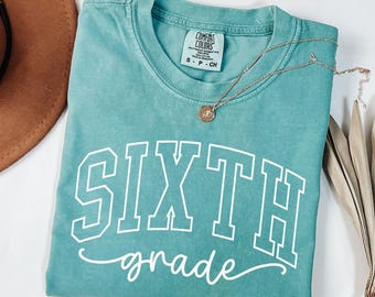 May include: Seafoam green t-shirt with the words "SIXTH grade" in white block letters and script. A gold necklace with a pendant rests on the shirt. The shirt is labelled "Comfort Colors" and is size S-P-CH.