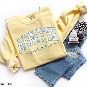 May include: A light yellow Auntie sweatshirt with the name "Sarah" in a script font. The word "AUNTIE" is in a blue floral design. The sweatshirt is paired with blue jeans and a small gift bag.