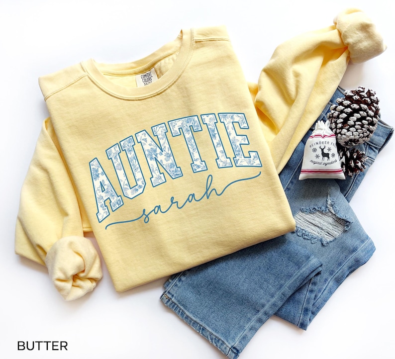 May include: A light yellow Auntie sweatshirt with the name "Sarah" in a script font. The word "AUNTIE" is in a blue floral design. The sweatshirt is paired with blue jeans and a small gift bag.