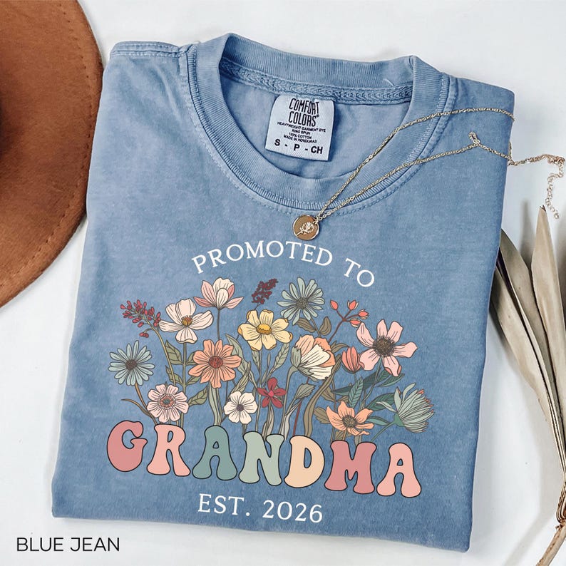 May include: Blue jean colored t-shirt with the words "PROMOTED TO GRANDMA EST. 2026" in a retro font. The shirt features a floral design in pastel colors. A gold necklace with a pendant is draped across the shirt.