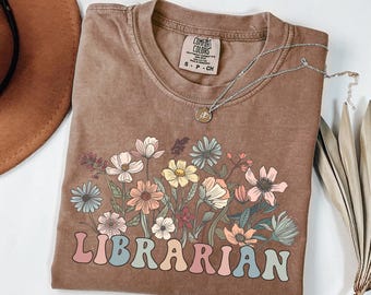 May include: Espresso-colored t-shirt with a floral design and the word "LIBRARIAN" in pastel colors. The shirt has a crew neck and short sleeves. A silver necklace with a pendant is draped across the shirt.
