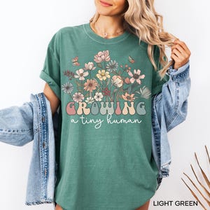 May include: Light green t-shirt with a floral design and the text "GROWING a tiny human" in a retro font. The shirt features a crew neck and short sleeves. The design includes various flowers and butterflies.