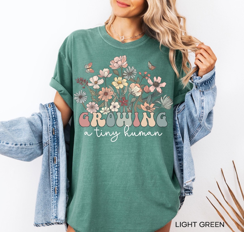 May include: Light green t-shirt with a floral design and the text "GROWING a tiny human" in a retro font. The shirt features a crew neck and short sleeves. The design includes various flowers and butterflies.