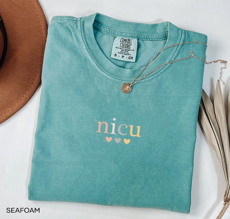 May include: Seafoam green Comfort Colors t-shirt with the word "nicu" in a gradient font and three hearts below. A gold necklace with a pendant rests on the shirt. The shirt is folded and has a tag that says "Comfort Colors".