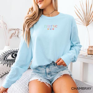 May include: Light blue long-sleeve shirt with the word "nurse" embroidered in pastel colors, with three hearts below. The shirt is paired with denim shorts. The word "CHAMBRAY" is printed in the bottom right corner.
