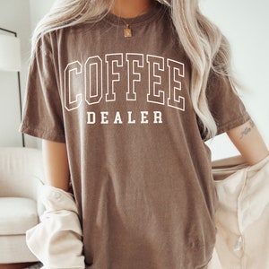 May include: Espresso-colored t-shirt with the text "COFFEE DEALER" in a white, collegiate-style font. The shirt has a relaxed fit and short sleeves. The word "ESPRESSO" is printed at the bottom left corner of the image.