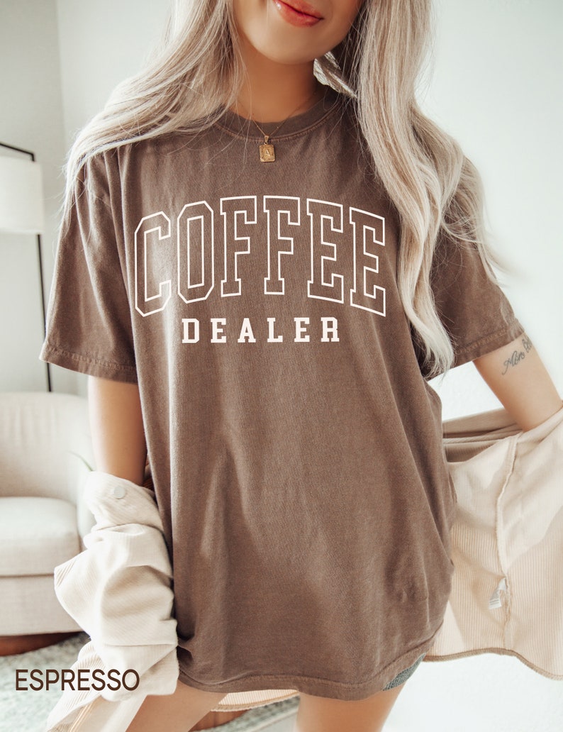 May include: Espresso-colored t-shirt with the text "COFFEE DEALER" in a white, collegiate-style font. The shirt has a relaxed fit and short sleeves. The word "ESPRESSO" is printed at the bottom left corner of the image.