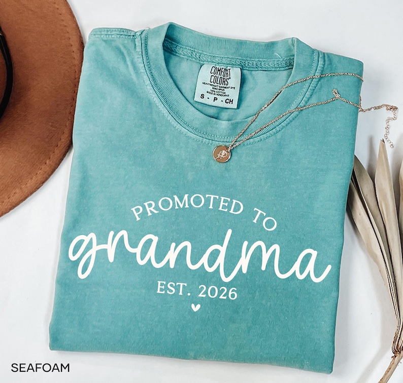 May include: Seafoam green t-shirt with the words "PROMOTED TO grandma EST. 2026" in white script. A gold necklace with a pendant rests on the shirt. The shirt is folded and placed on a white surface.