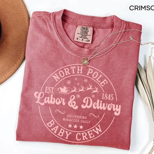 May include: Crimson-colored t-shirt with a crew neck and a circular graphic that reads "North Pole Labor & Delivery Baby Crew" with a sleigh and reindeer design. A gold necklace rests on the shirt. The shirt is next to a brown hat and dried leaves.
