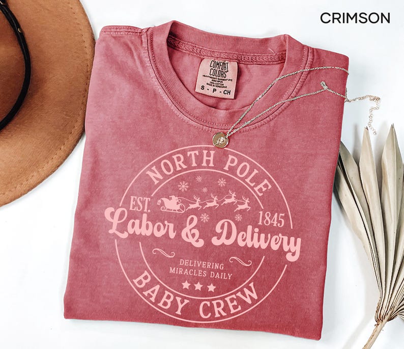 May include: Crimson-colored t-shirt with a crew neck and a circular graphic that reads "North Pole Labor & Delivery Baby Crew" with a sleigh and reindeer design. A gold necklace rests on the shirt. The shirt is next to a brown hat and dried leaves.