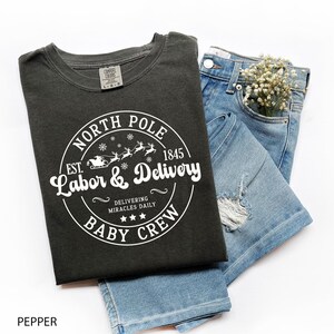 May include: A dark gray t-shirt with the text "NORTH POLE LABOR & DELIVERY BABY CREW" in a circular design. The shirt is paired with blue denim shorts. The text includes a Santa sleigh and reindeer graphic.