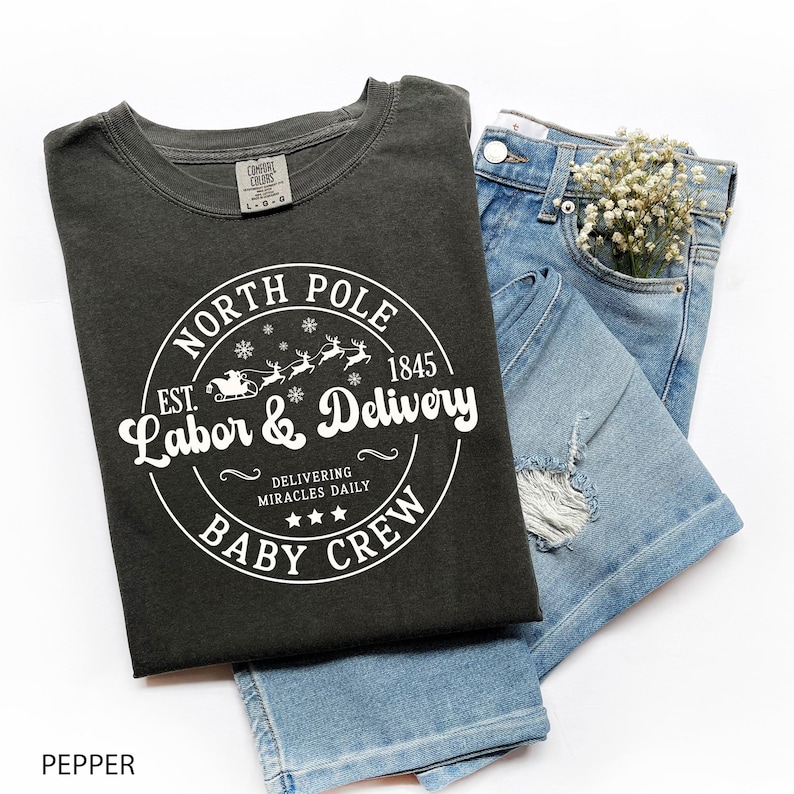 May include: A dark gray t-shirt with the text "NORTH POLE LABOR & DELIVERY BABY CREW" in a circular design. The shirt is paired with blue denim shorts. The text includes a Santa sleigh and reindeer graphic.