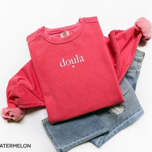 May include: A red sweatshirt with the word "doula" printed in white, with a small heart beneath the word. The sweatshirt is folded over a pair of blue denim jeans. The word "WATERMELON" is printed at the bottom.