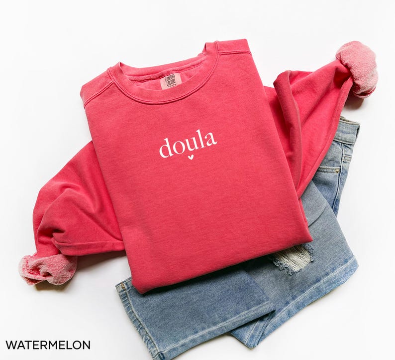 May include: A red sweatshirt with the word "doula" printed in white, with a small heart beneath the word. The sweatshirt is folded over a pair of blue denim jeans. The word "WATERMELON" is printed at the bottom.