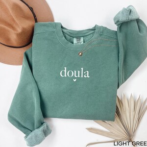 May include: A light green sweatshirt with the word "doula" printed in white, with a small heart symbol underneath. The sweatshirt has a crew neck and long sleeves with rolled cuffs. A gold necklace is visible near the neckline.
