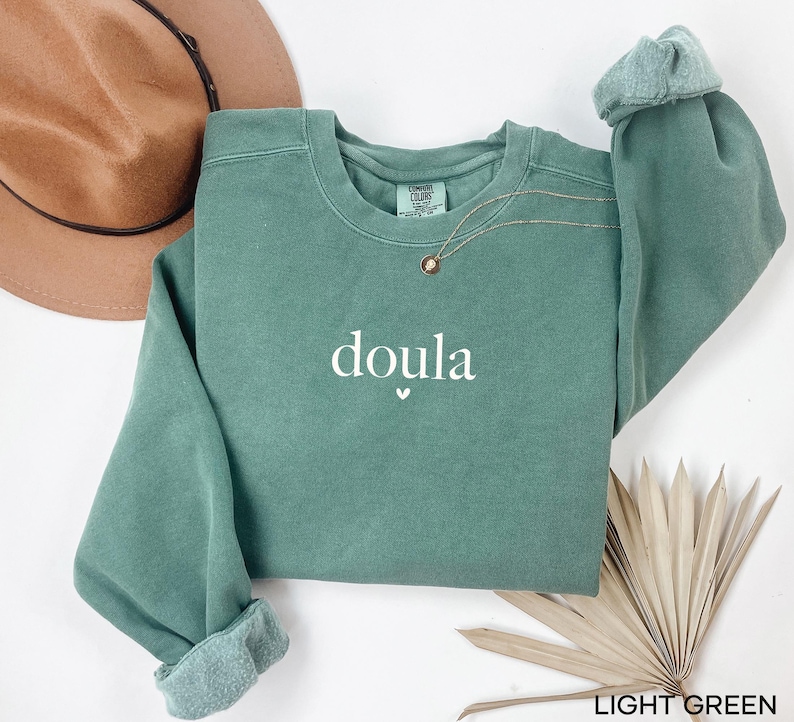 May include: A light green sweatshirt with the word "doula" printed in white, with a small heart symbol underneath. The sweatshirt has a crew neck and long sleeves with rolled cuffs. A gold necklace is visible near the neckline.