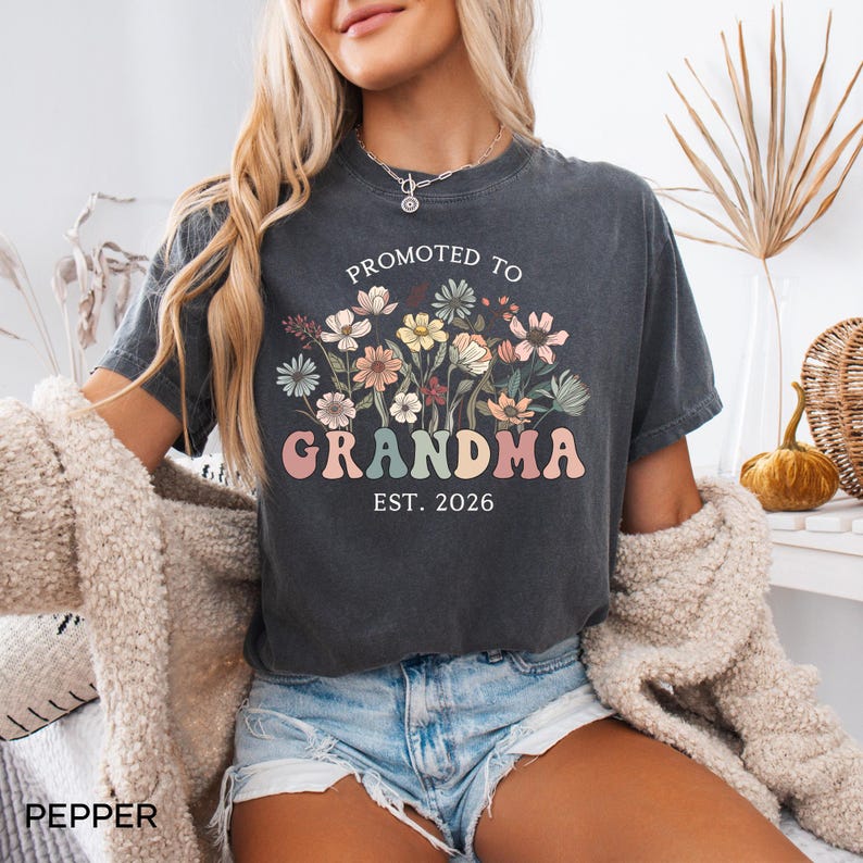 May include: Dark gray t-shirt with the words "PROMOTED TO GRANDMA EST. 2026" in a floral design. The design features pastel-colored flowers and text. The shirt is worn with denim shorts and a cream-colored cardigan.