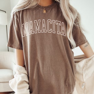 K&ouml;nnte beinhalten: Ein braunes Oversize-T-Shirt mit dem Text "MAMACITA" in wei&szlig;en umrandeten Buchstaben.