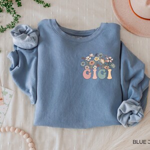 以下が含まれることがあります： 花柄のデザインとカラフルな文字で「GIGI」と書かれた青いスウェットシャツです。