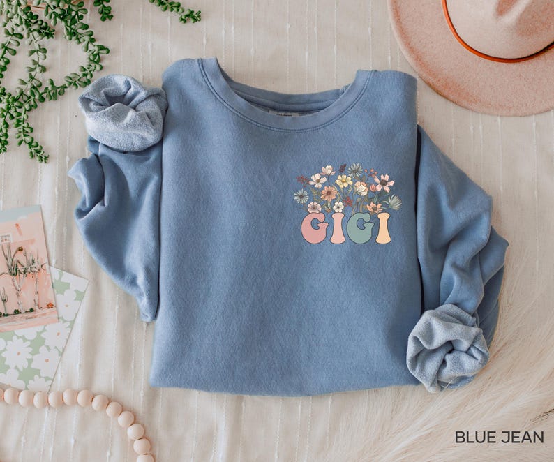 以下が含まれることがあります： 花柄のデザインとカラフルな文字で「GIGI」と書かれた青いスウェットシャツです。