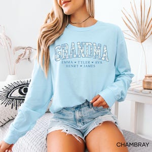 May include: A light blue long-sleeve sweatshirt with the word "GRANDMA" in a floral design. Below the word are the names Emma, Tyler, Ava, Henry, and James. The word "CHAMBRAY" is printed at the bottom right of the image.