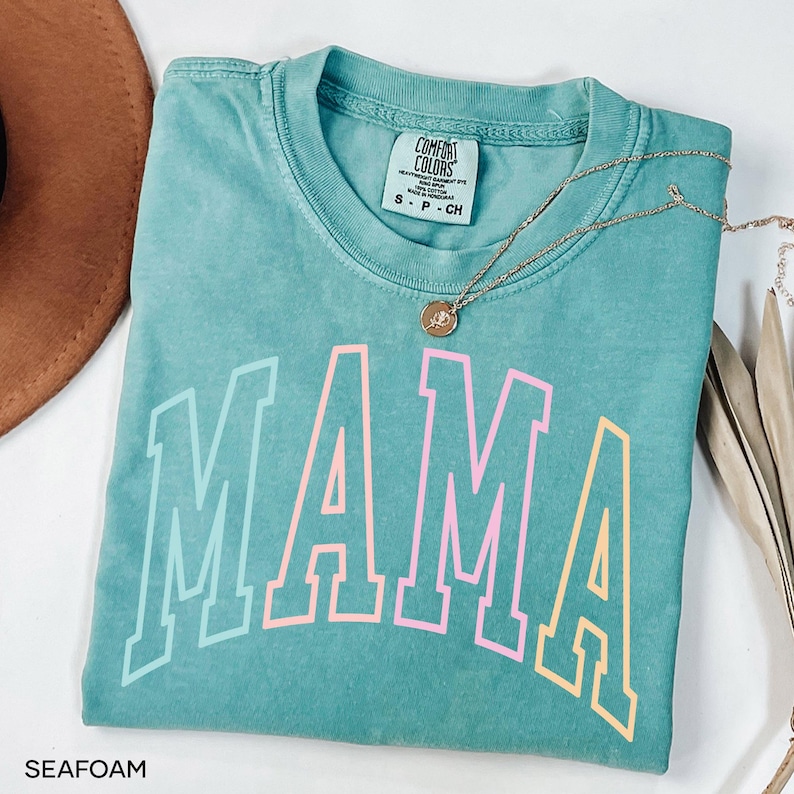 May include: Seafoam green t-shirt with the word "MAMA" in rainbow colors. The shirt has a distressed look and is made of a soft, comfortable fabric.