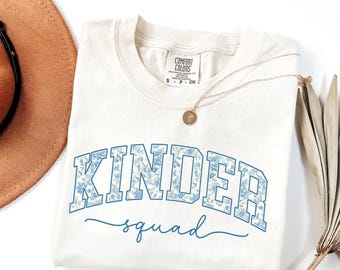 May include: Ivory t-shirt featuring the words "KINDER squad" in a blue floral design. The top has a crew neck and short sleeves. A gold necklace with a pendant is draped across the shirt.