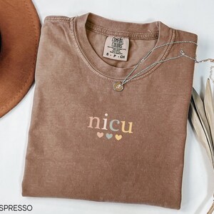 May include: Espresso-colored t-shirt with the word "nicu" in pastel colors and three small hearts. A silver necklace with a pendant rests on the shirt. The shirt is folded and has a tag that says "Comfort Colors".