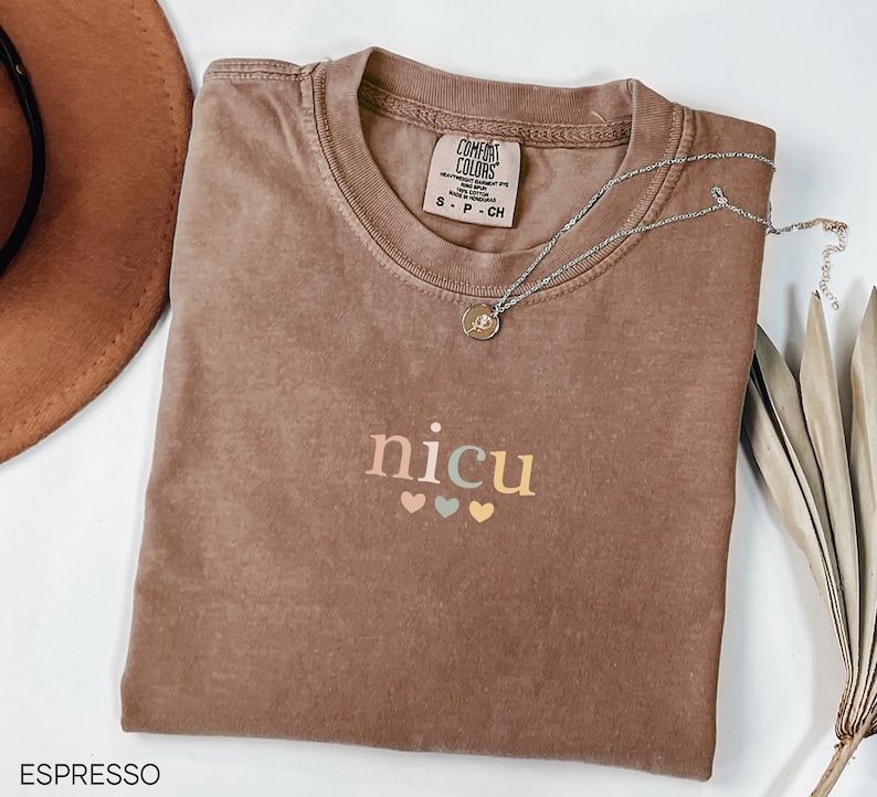 May include: Espresso-colored t-shirt with the word "nicu" in pastel colors and three small hearts. A silver necklace with a pendant rests on the shirt. The shirt is folded and has a tag that says "Comfort Colors".