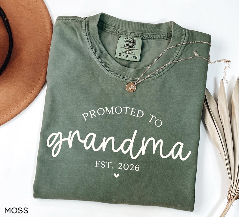 May include: A moss green t-shirt with the words "Promoted to grandma" in white script, along with "Est. 2026" and a small heart. A gold necklace with a pendant rests on the shirt. The shirt is folded.