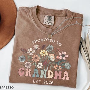 May include: Espresso-colored t-shirt with the text "PROMOTED TO GRANDMA EST. 2026" in a colorful, floral design. The shirt has a crew neck and short sleeves. A silver necklace with a pendant is draped across the shirt.