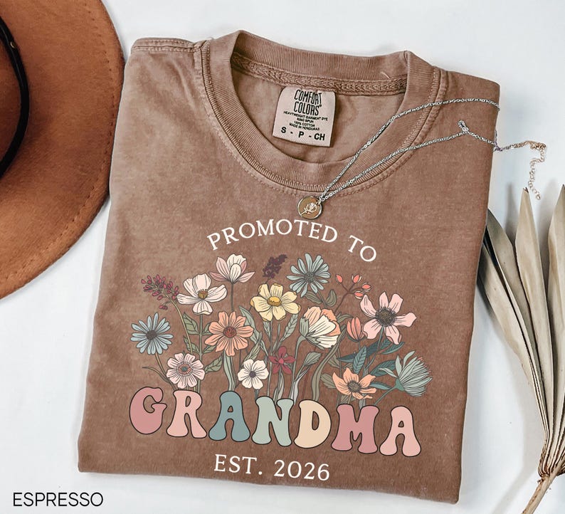 May include: Espresso-colored t-shirt with the text "PROMOTED TO GRANDMA EST. 2026" in a colorful, floral design. The shirt has a crew neck and short sleeves. A silver necklace with a pendant is draped across the shirt.