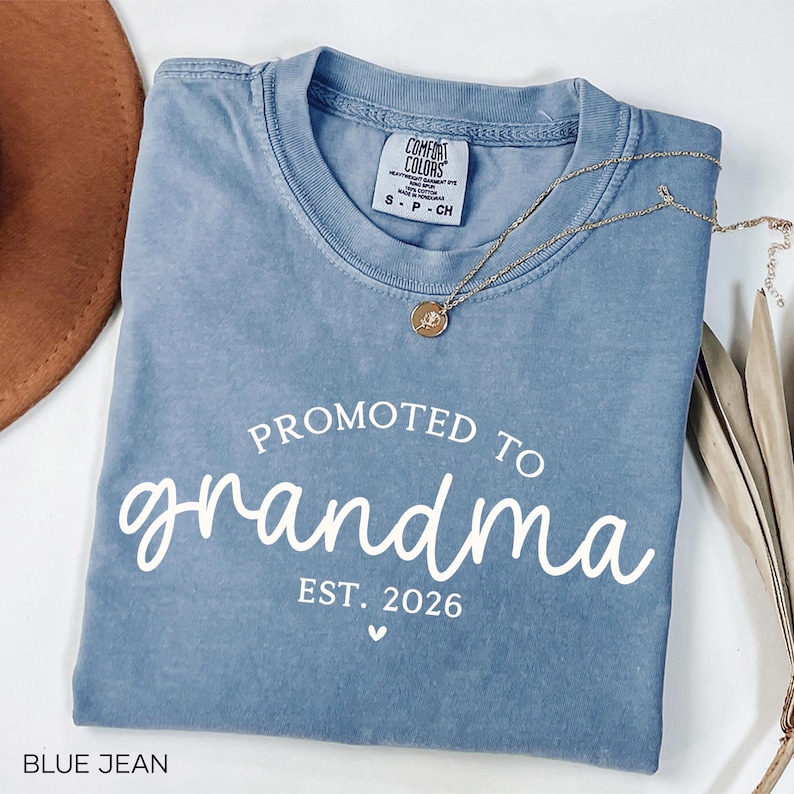May include: A blue jean Comfort Colors t-shirt with the words "Promoted to Grandma" and "Est. 2026" printed in white. A gold necklace with a pendant is draped across the shirt. A brown hat is visible in the upper left corner.