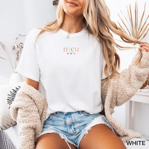 May include: White t-shirt with the word "NICU" in a soft, pastel font, with three small hearts underneath. The shirt is paired with denim shorts and a beige cardigan. The word "WHITE" is visible in the bottom right corner.