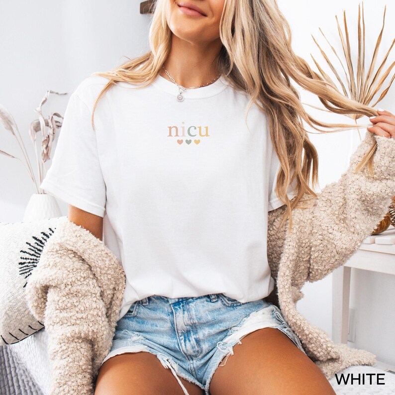 May include: White t-shirt with the word "NICU" in a soft, pastel font, with three small hearts underneath. The shirt is paired with denim shorts and a beige cardigan. The word "WHITE" is visible in the bottom right corner.