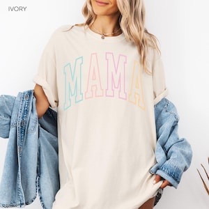 May include: A light beige t-shirt with the word "MAMA" in rainbow colors. The letters are outlined in black and have a varsity style font.